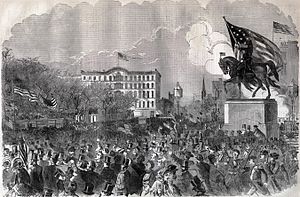 Crowd surrounding an equestrian statue toped by a huge U.S. flag.